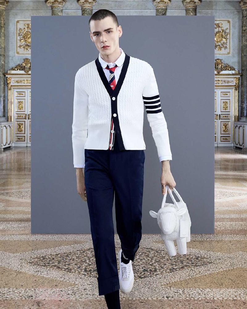 Fashion shot of a slender adult male wearing a white cardigan, dress shirt, and dress pants holding a sheep-shaped handbag.