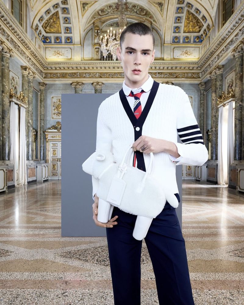 Fashion shot of a young adult male in a white cardigan holding a plush, white sheep-shaped bag in a baroque-style interior space.