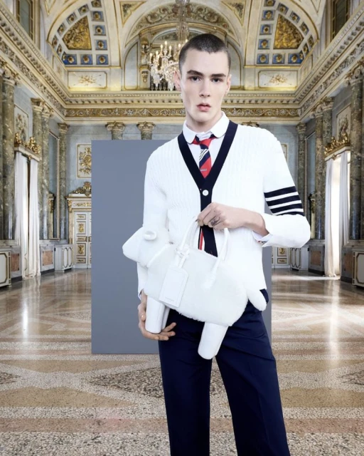 Fashion shot of a young adult male in a white cardigan holding a plush, white sheep-shaped bag in a baroque-style interior space.