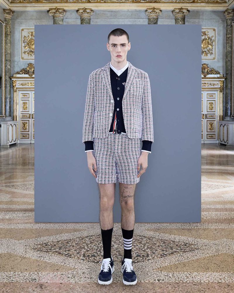 Fashion shot of a young adult male model wearing a tweed suit with navy socks and trainers, standing in front of grey backdrop in palace.