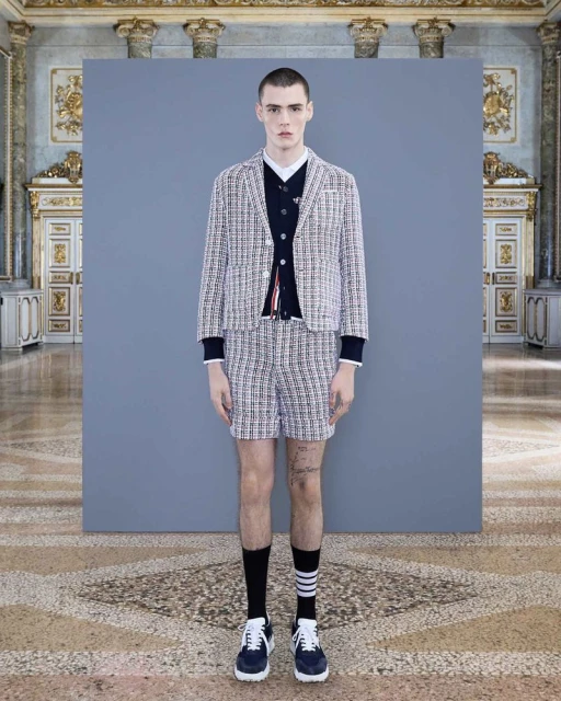 Fashion shot of a young adult male model wearing a tweed suit with navy socks and trainers, standing in front of grey backdrop in palace.