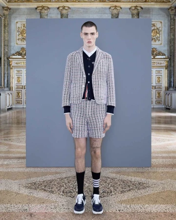 Fashion shot of a young adult male model wearing a tweed suit with navy socks and trainers, standing in front of grey backdrop in palace.