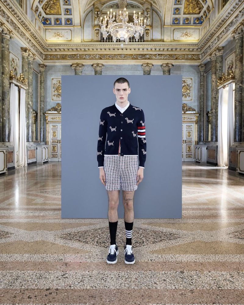 Male fashion model in a formal cardigan with dog motifs, shorts, and sneakers, posing indoors in an opulent palace-like setting.