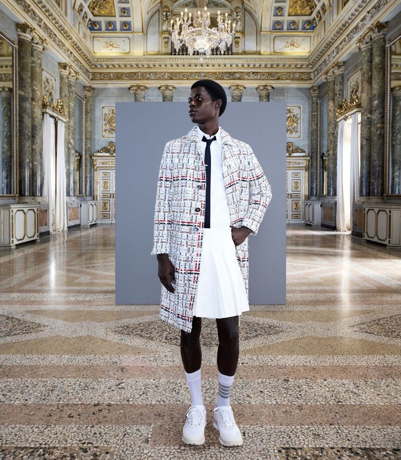 Fashion shot of a black male model wearing a white pleated skirt and patterned coat posing in an ornate building with a tiled floor.