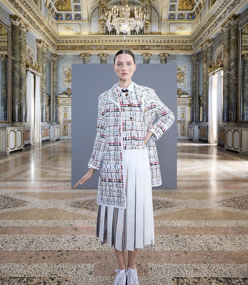 Fashion shot of a woman in a white tweed jacket and pleated skirt, standing in an ornate ballroom with high ceilings and marble floors.