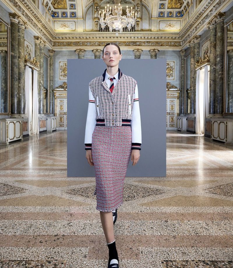 Fashion shot of a female model in a patterned skirt and vest, standing in a grand hall with a high ceiling and stone floors.