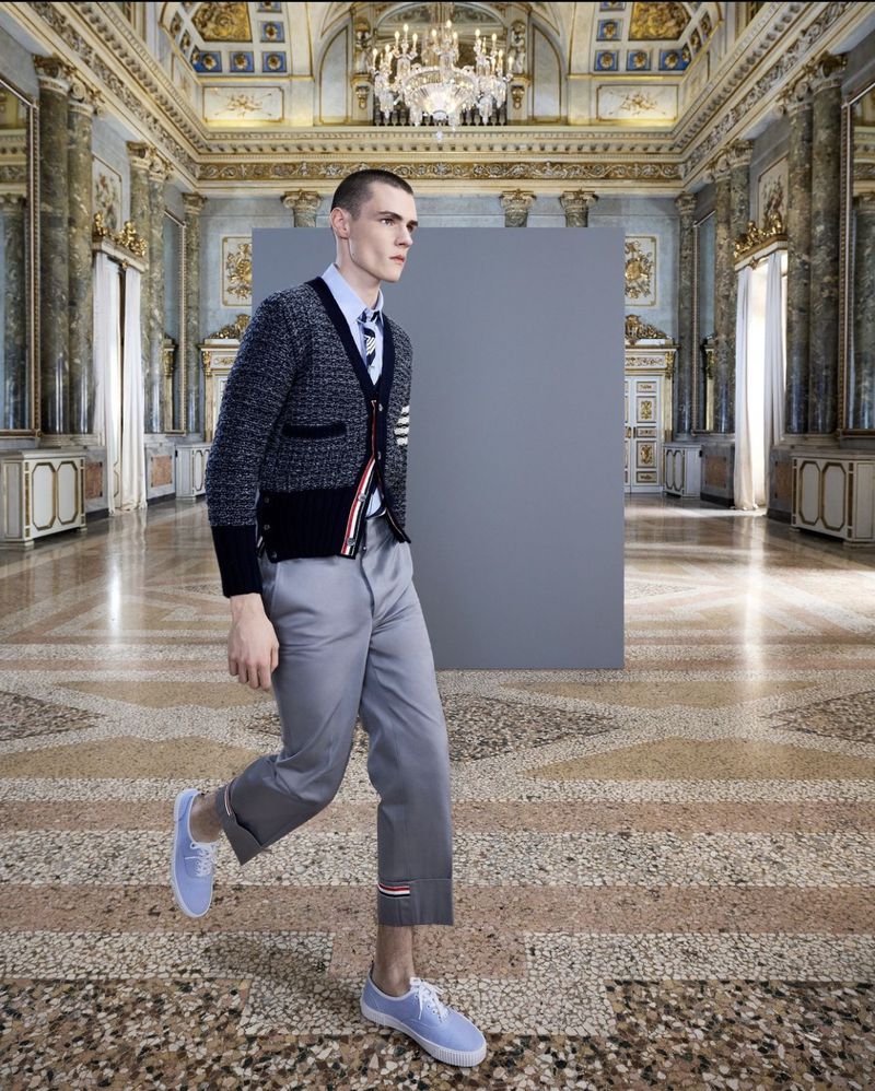 Man in grey wool cardigan, blue dress shirt, grey capri pants, and blue sneakers walking on ornate stone floor with grey backdrop.
