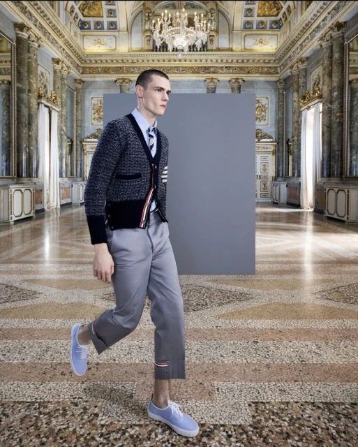 Man in grey wool cardigan, blue dress shirt, grey capri pants, and blue sneakers walking on ornate stone floor with grey backdrop.
