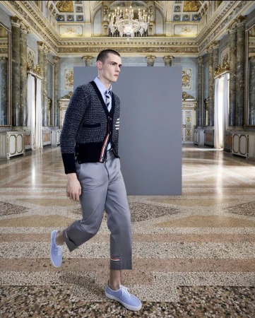 Man in grey wool cardigan, blue dress shirt, grey capri pants, and blue sneakers walking on ornate stone floor with grey backdrop.