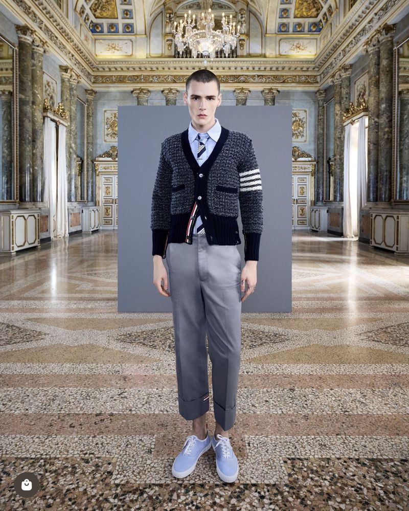 Male model in cardigan, shirt, and trousers poses in a grand hall with a patterned stone floor and ornate wall and ceiling details.