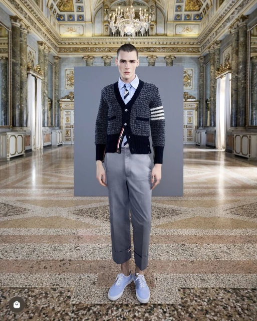 Male model in cardigan, shirt, and trousers poses in a grand hall with a patterned stone floor and ornate wall and ceiling details.