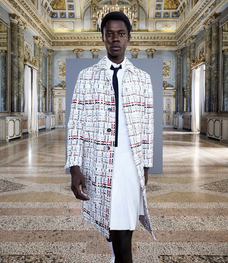 Fashion shot of a slender Black male model wearing a patterned coat, white shirt, and pleated skirt in an ornate marble hall.