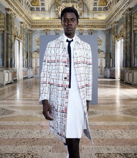 Fashion shot of a slender Black male model wearing a patterned coat, white shirt, and pleated skirt in an ornate marble hall.