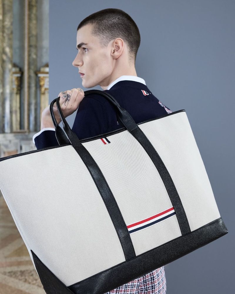 Close-up of an adult male carrying a large off-white tote bag with black leather straps and trim in a brightly lit indoor setting.