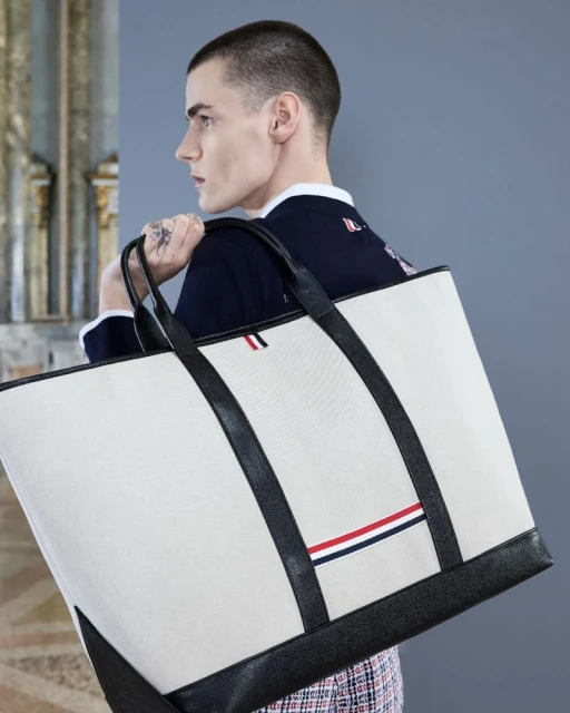 Close-up of an adult male carrying a large off-white tote bag with black leather straps and trim in a brightly lit indoor setting.