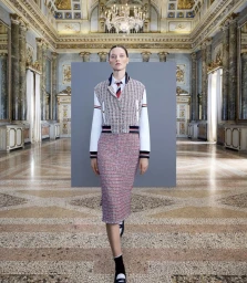 Model wearing a tweed skirt suit and striped tie standing in an ornate hall with marble columns and a crystal chandelier.