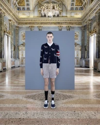 Model standing centrally in a navy cardigan and patterned shorts against a grey backdrop inside an ornate hall.