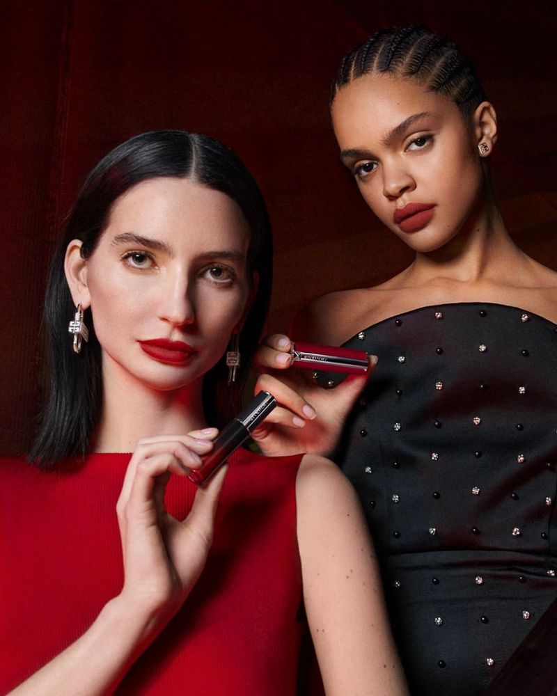 Two elegant women pose with dark red lipstick in a close-up beauty shot with a dark red and black background.