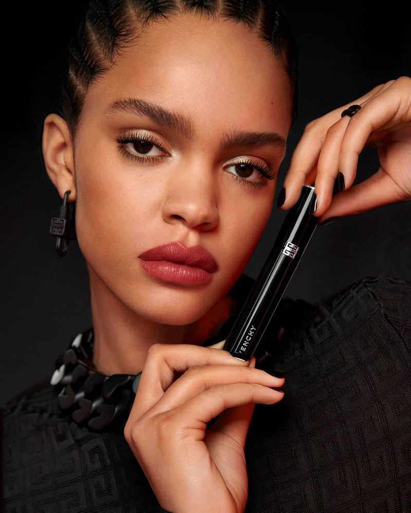 Close-up of a woman holding Givenchy cosmetics, showcasing flawless skin, elegant black top, and jewelry against a dark backdrop.