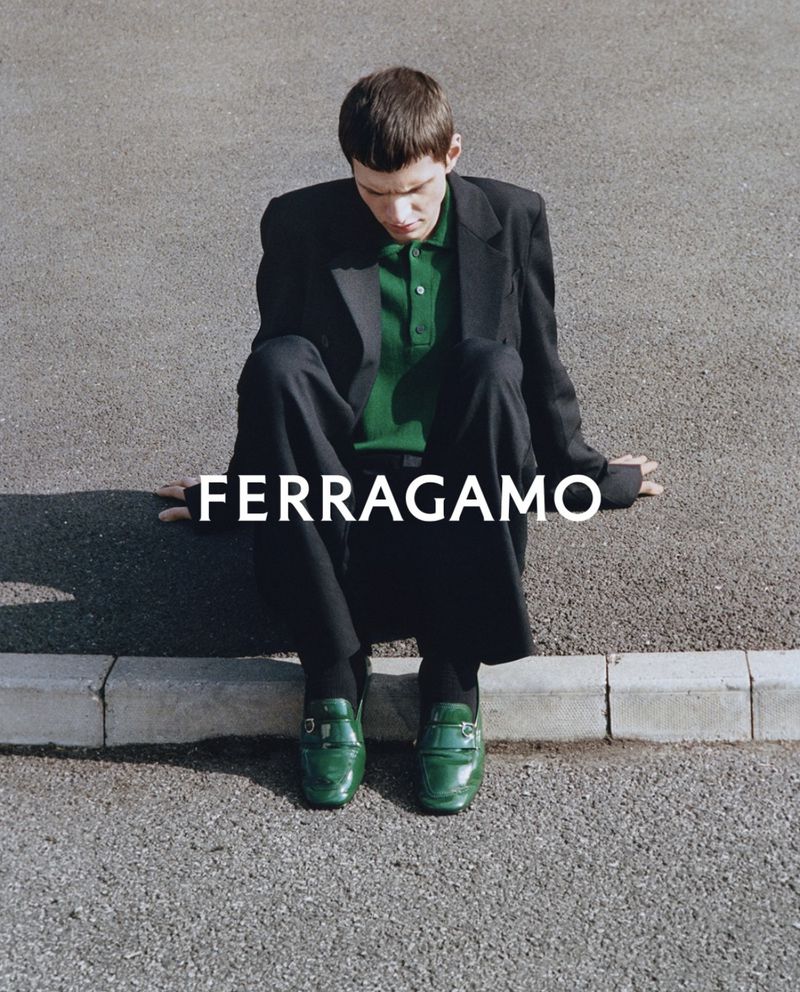 A person in a dark coat and bright green loafers sitting on the edge of pavement near a road with the word FERRAGAMO in white letters.