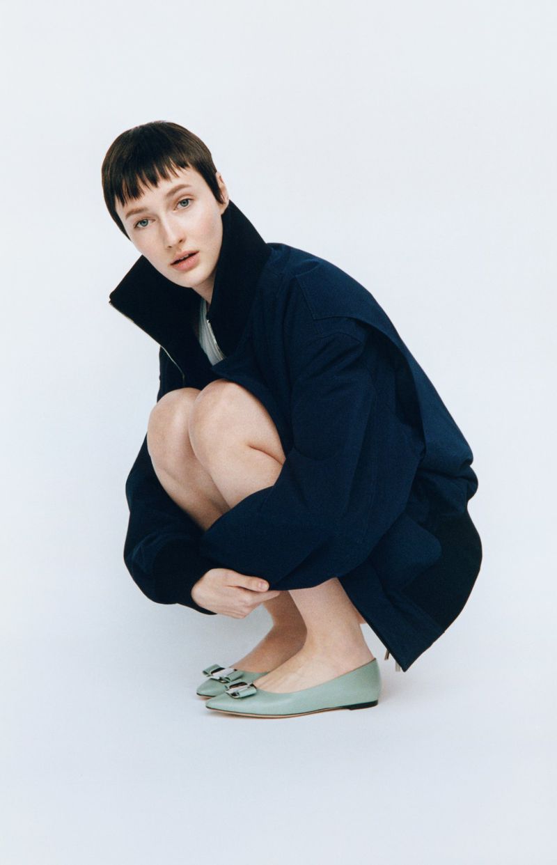 Woman in crouching pose wearing a navy jacket and light green flats against a white backdrop in a fashion studio shot.
