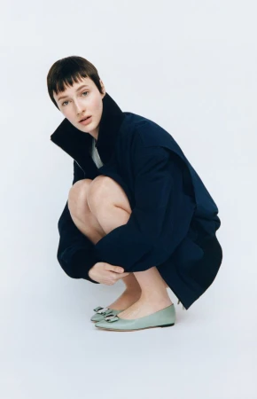 Woman in crouching pose wearing a navy jacket and light green flats against a white backdrop in a fashion studio shot.