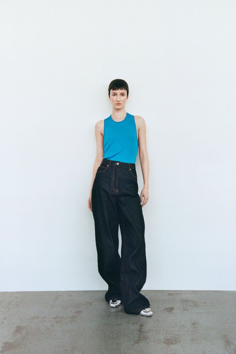 A full shot features a woman with short brown hair wearing a turquoise tank top and wide-leg denim jeans against a neutral background.
