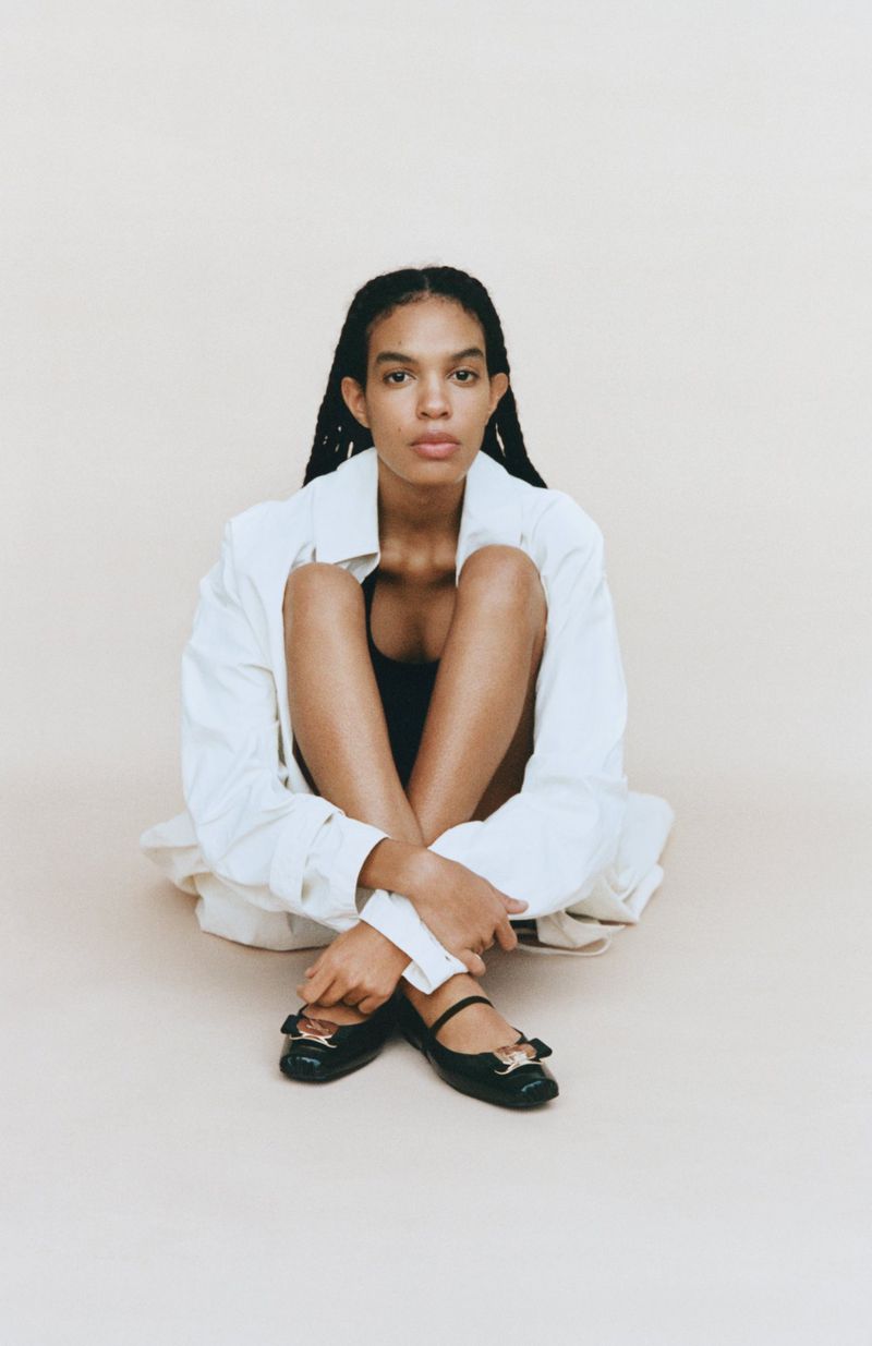 A young adult woman sits on the floor wearing a white button-down shirt, black top, and black ballet flats in a studio setting.