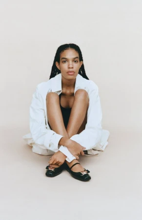A young adult woman sits on the floor wearing a white button-down shirt, black top, and black ballet flats in a studio setting.