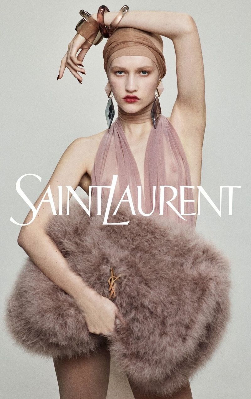 Saint Laurent fashion ad: a young woman in a sheer top and headscarf holds a feathery bag, accessorized with cuffs and earrings.