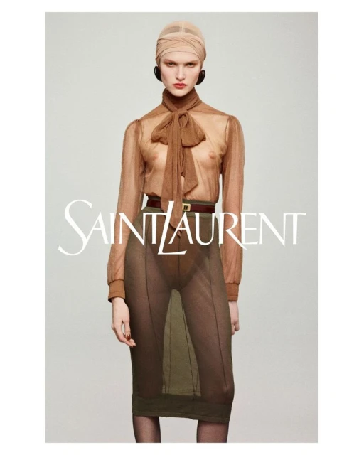 Fashion editorial shot: Brunette model in sheer brown blouse, olive skirt, and head wrap poses against light gray backdrop with brand logo.