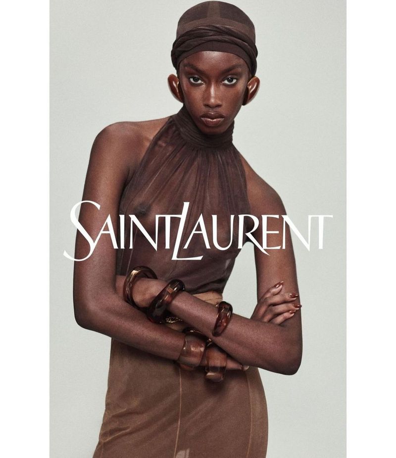 Fashion portrait of a woman wearing a sheer brown halter top, matching head wrap, skirt, and bracelets against a neutral backdrop.