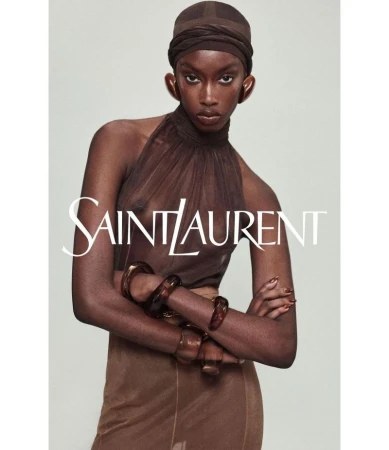 Fashion portrait of a woman wearing a sheer brown halter top, matching head wrap, skirt, and bracelets against a neutral backdrop.