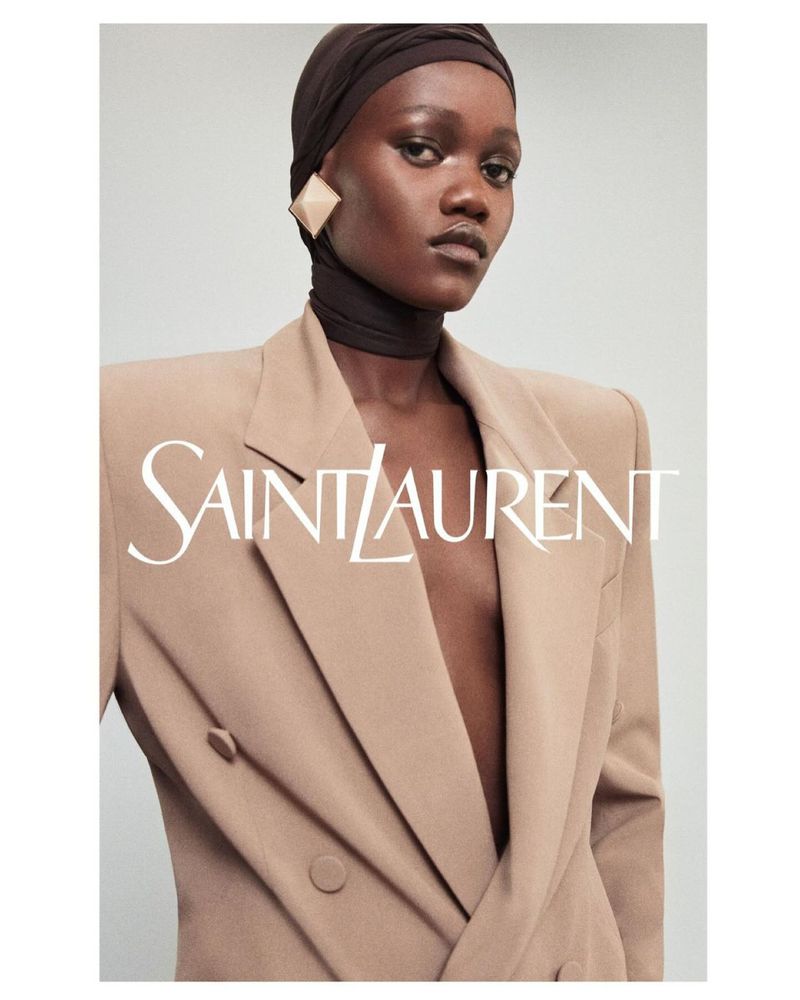 Elegant portrait of a Black woman wearing a turban and double-breasted suit, showcasing diamond earrings and the Saint Laurent logo.