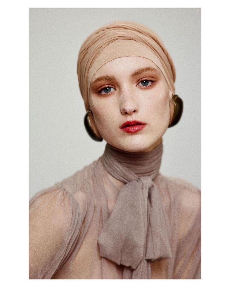 Close-up portrait of a woman with peach-tone head wrap, sheer top with bow, and large earrings against a neutral background.