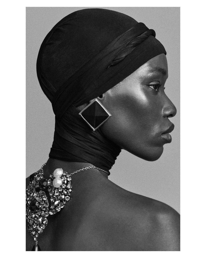 Profile portrait of an African woman with black head wrap, jewelry, and dark skin, shot in a photo studio with gray background.
