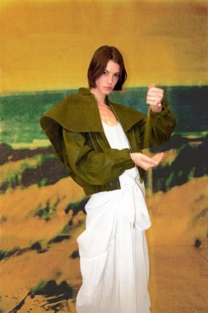 A young woman with short brunette hair is seen holding sand in her hands, wearing a green jacket and white dress against a scenic backdrop.