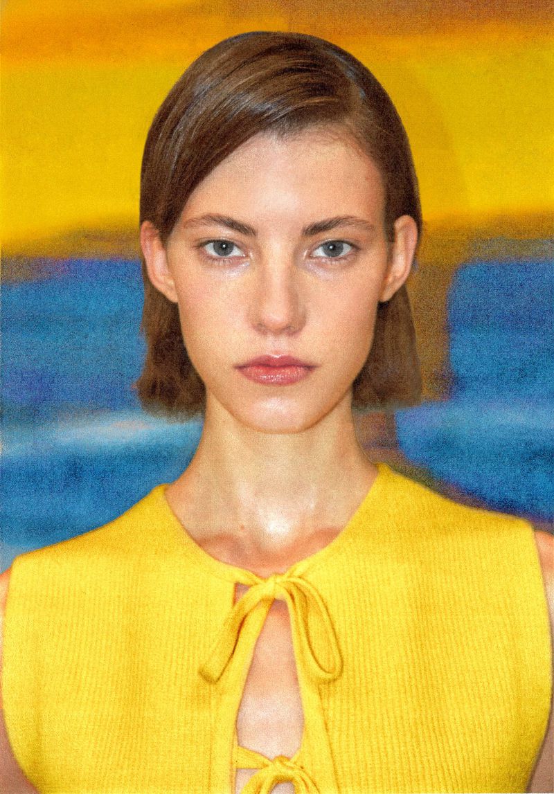 Close-up portrait of a young adult woman with short brown hair wearing a yellow top against a painted yellow and blue background.