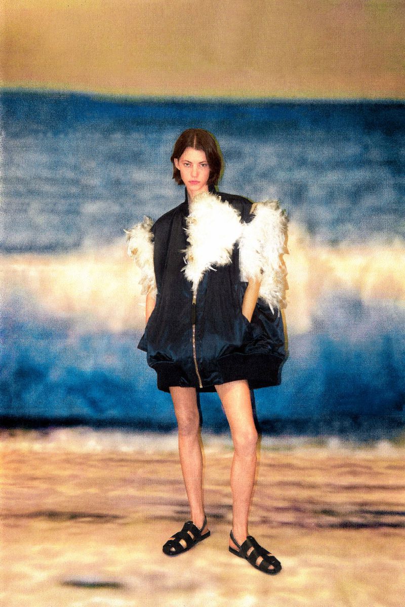 Fashion shot of a young woman in a dark jacket with white feather trim, black skirt, and sandals, posing on a beach with ocean backdrop.