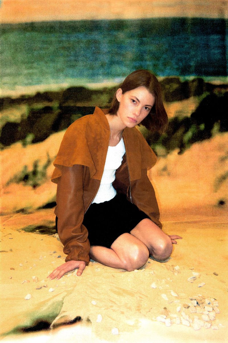 Young woman kneels on sand in a tan jacket, white top, and black skirt against a blurry backdrop of beach and ocean scenery.