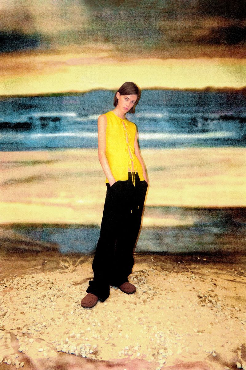 Young adult poses in yellow vest and dark pants against a painted backdrop of beach, sky, and horizon line on a gravelly ground.