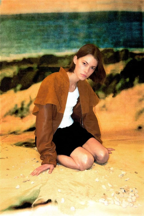 Model kneeling on sandy ground wearing a brown leather jacket, white top, and black skirt against a painted coastal backdrop.
