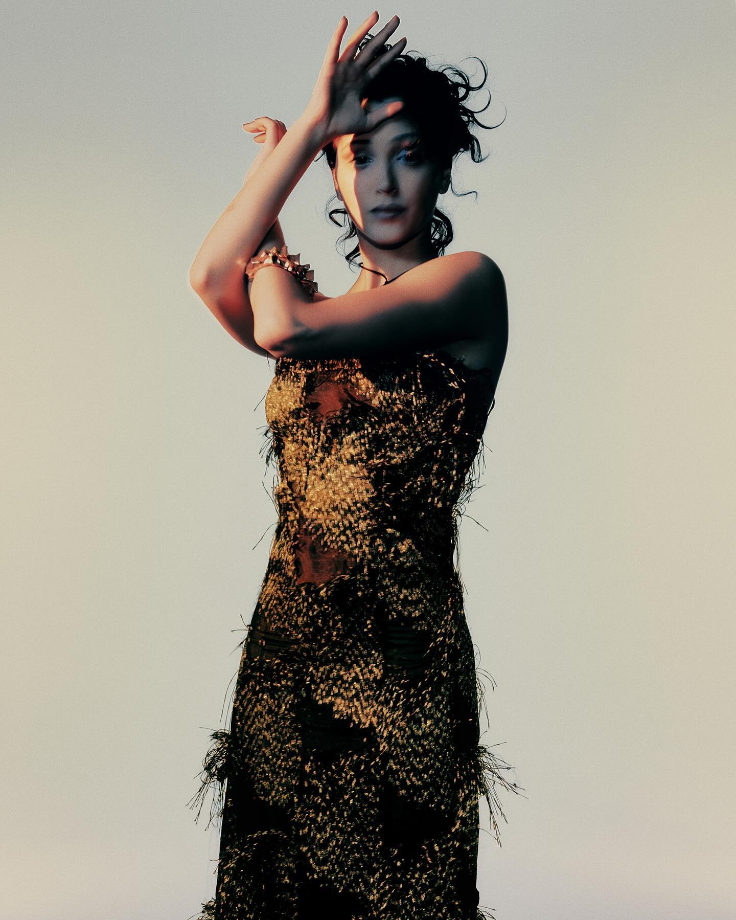 Woman posing in a textured black and gold dress with her arms raised near her face against a neutral background.