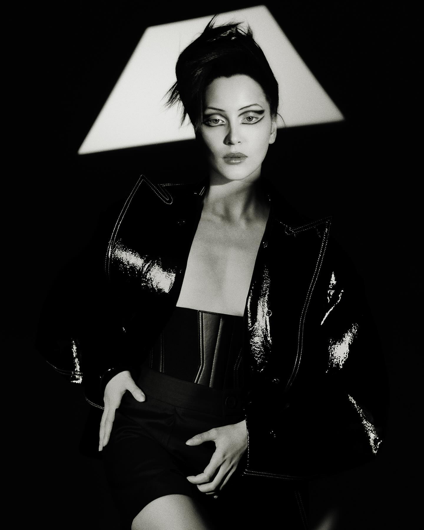 Monochrome portrait of an adult female model in a black patent leather jacket with dramatic eye makeup posing in a photo studio.