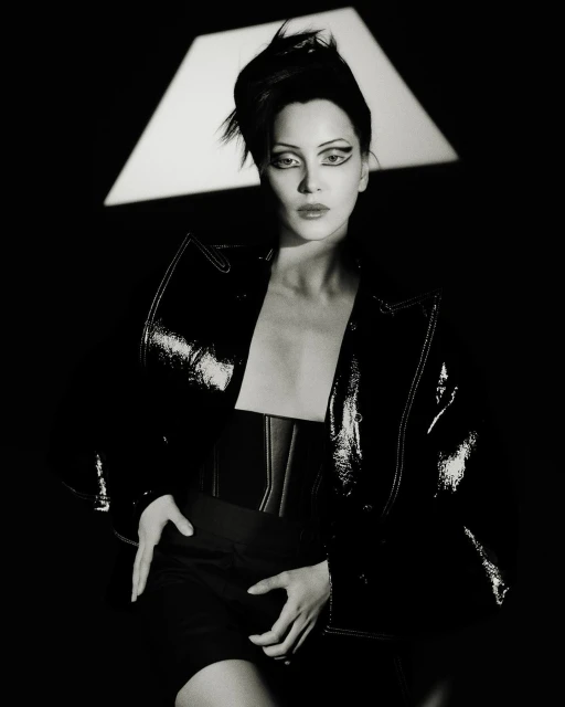 Monochrome portrait of an adult female model in a black patent leather jacket with dramatic eye makeup posing in a photo studio.