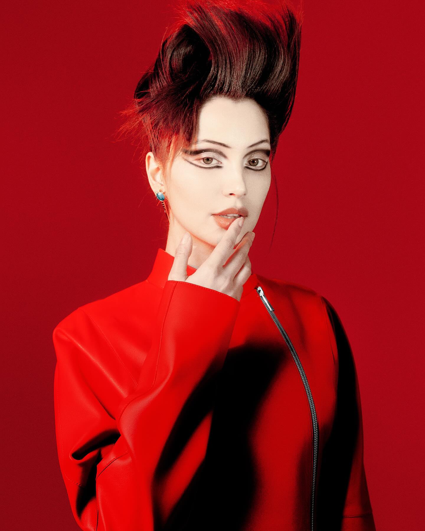A striking portrait of a woman with dramatic makeup and an avant-garde hairstyle, wearing a red jacket against a red backdrop in studio.