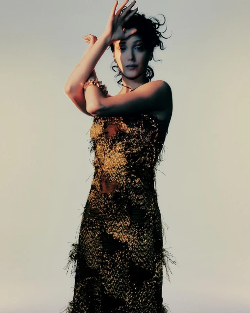 A woman poses in a gold fringed gown against a neutral background, hands raised to her face, and wearing a gold bracelet.