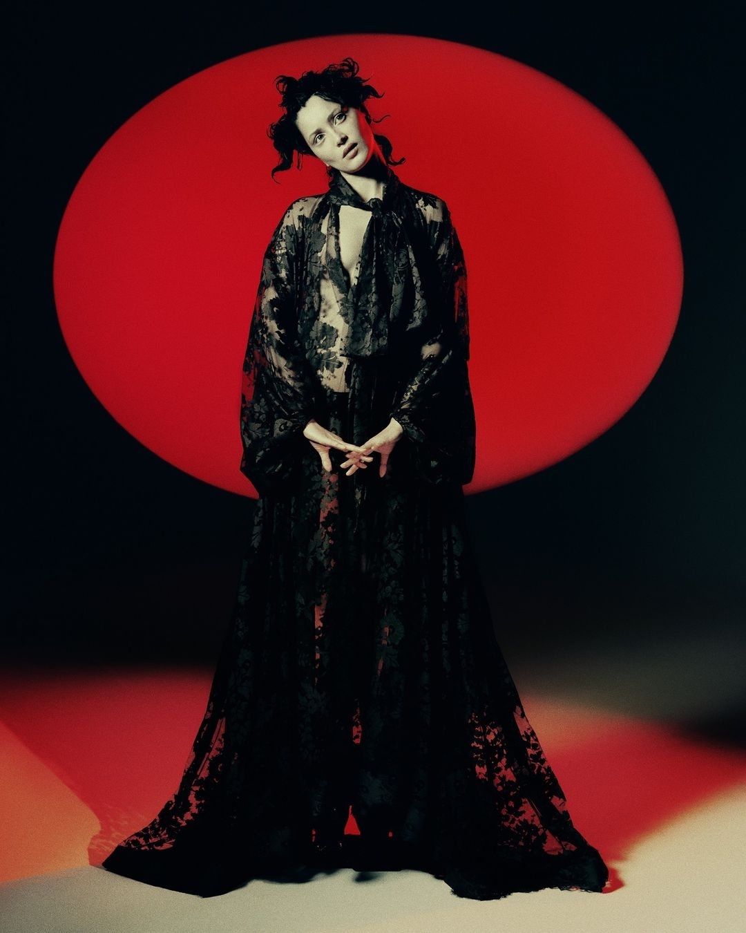 A fair-skinned adult woman poses in a floor-length black lace dress with a red circle in the background against a dark backdrop.