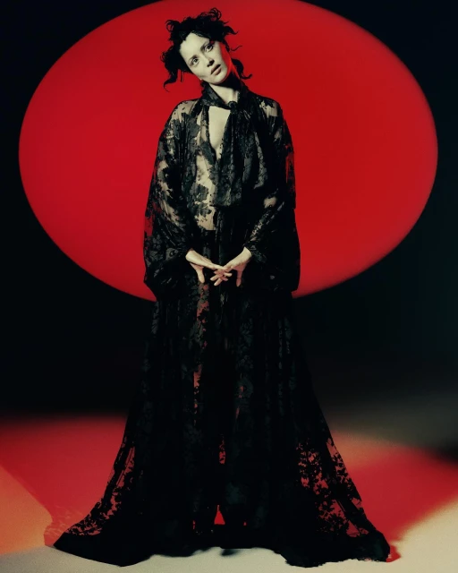 A fair-skinned woman with dark hair wearing a sheer black lace robe stands in front of a large red circle against a black background.