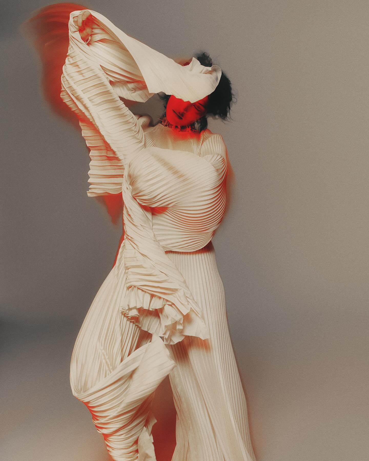 Model in pleated cream clothing moving dynamically with blurred red light trails against a grey studio backdrop.